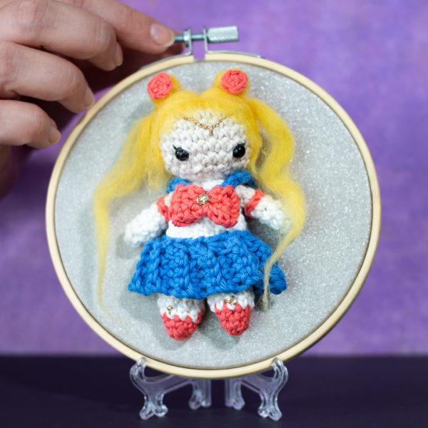 Tableau avec un amigurumi, figurine en crochet, représentant Sailor moon, guerrière de la lune, tenu par une main créée et réalisée par le Rat et l'Ours.