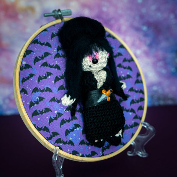 Tableau avec un amigurumi, figurine en crochet, représentant Elvira, maitresse des ténèbres, de profil, imaginé et confectionné à la main par le Rat et l'Ours.