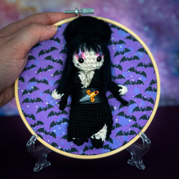 Tableau avec un amigurumi, figurine en crochet, représentant Elvira, maitresse des ténèbres, tenue en main imaginé et confectionné à la main par le Rat et l'Ours.