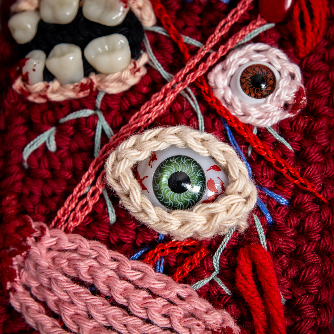 Détails d'un tableau en crochet sur le thème du body horror, avec des yeux, des dents, et des muscles comme un écorché… Œuvre imaginée et confectionnée à la main par les Mignonstres, une marque le Rat et l’Ours.