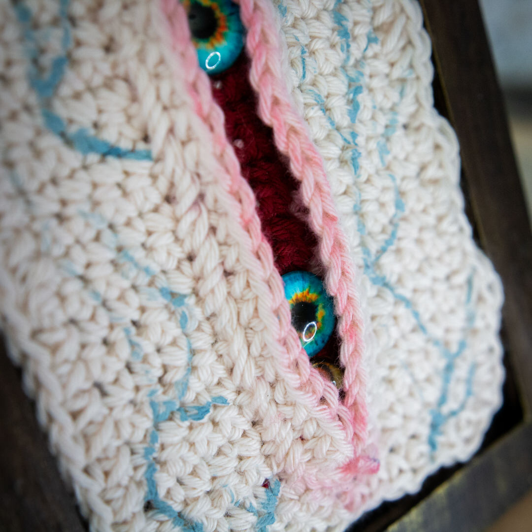 Détail d'un tableau en crochet sur le thème du body horror, avec une immense blessure qui a des yeux à l'intéreiru. Œuvre imaginée et confectionnée à la main par les Mignonstres, une marque le Rat et l’Ours.