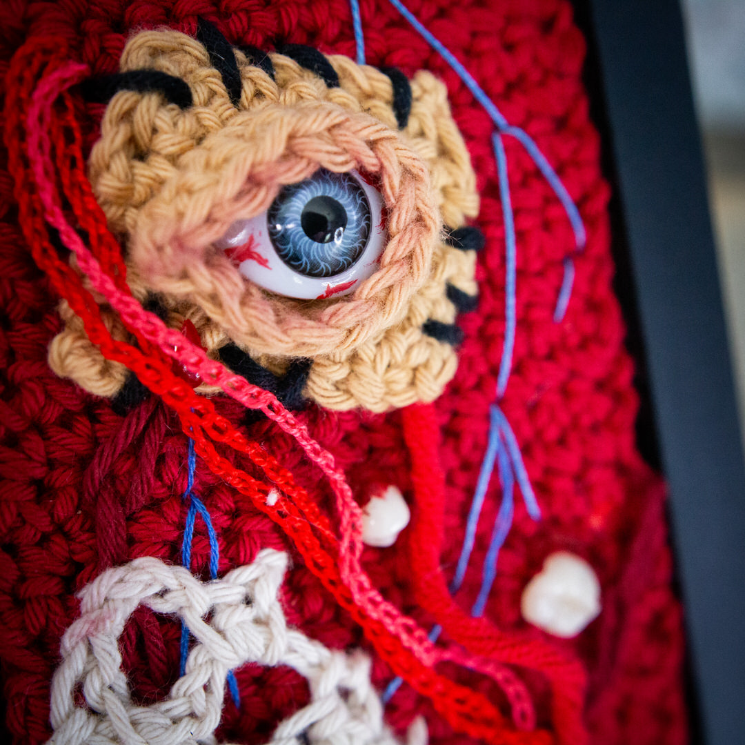 Détail d'un tableau en crochet sur le thème du body horror, avec des muscles à vifs, des dents, des veines et un oeil cousu. Œuvre imaginée et confectionnée à la main par les Mignonstres, une marque le Rat et l’Ours.