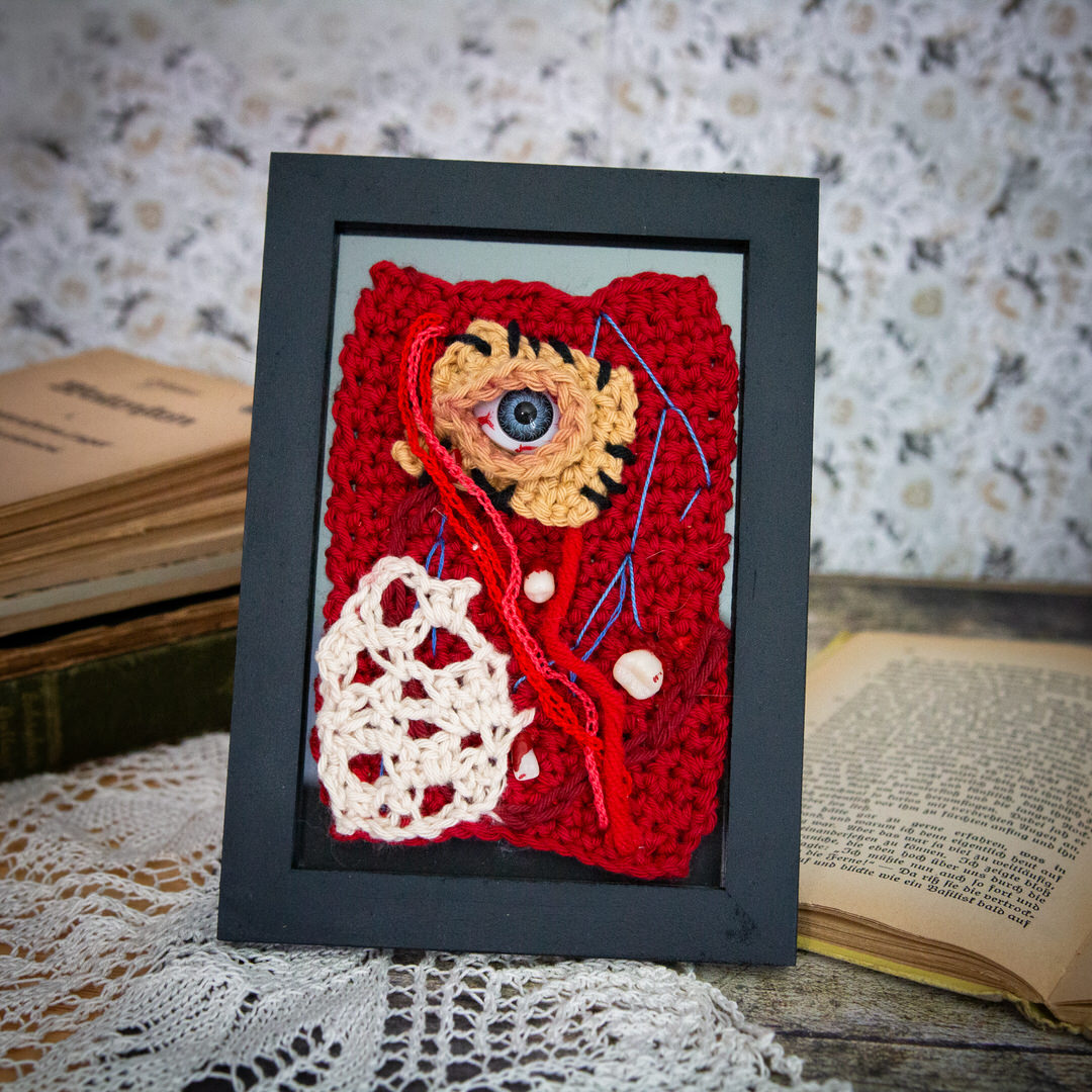 Tableau en crochet sur le thème du body horror, avec des muscles à vifs, des dents, des veines et un oeil cousu. Œuvre imaginée et confectionnée à la main par les Mignonstres, une marque le Rat et l’Ours.